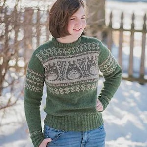 Könnte beinhalten: Grüner Strickpullover mit einem Totoro-Design in Grau- und Cremetönen. Der Pullover hat einen gerippten Saum und Bündchen sowie einen Rundhalsausschnitt. Die Person trägt blaue Jeans.