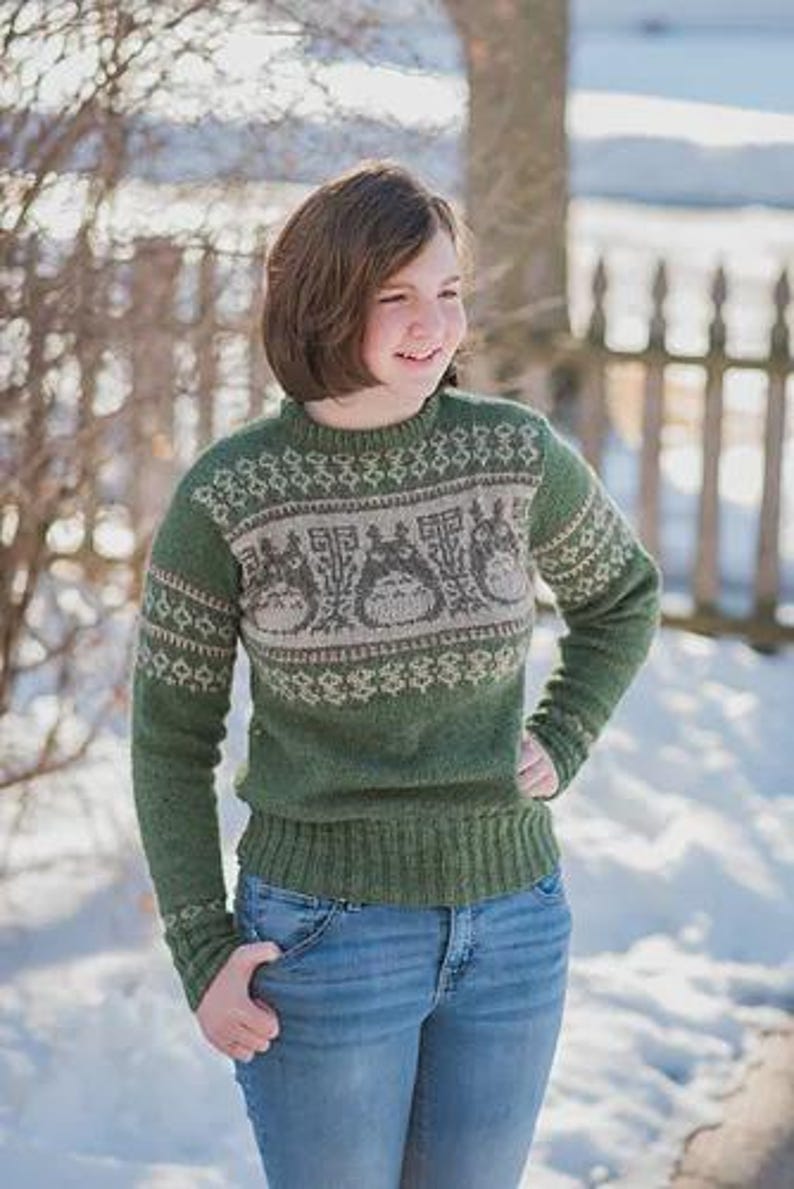 May include: Green knit sweater with a Totoro design in shades of gray and cream. The sweater has a ribbed hem and cuffs, and a crew neck. The person is wearing blue jeans.