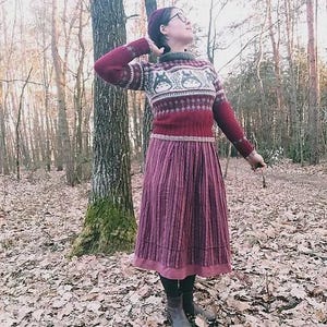 May include: A person wearing a burgundy sweater with a white and grey pattern, a burgundy skirt, and a burgundy beret. The person is standing in a forest with fallen leaves on the ground. The sweater has long sleeves and a high neck.
