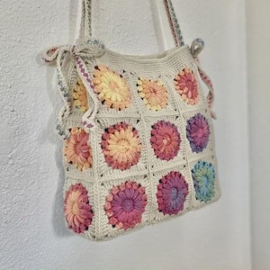 May include: A cream-colored crochet tote bag with a floral pattern. The bag features square panels with colorful, daisy-like flowers in shades of pink, yellow, and purple. The straps are also crocheted and tied with bows.