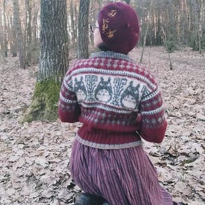 May include: A burgundy beret with gold embroidery and a red and gray sweater featuring a pattern of cartoon characters. The person is wearing a long, pleated, purple skirt and is kneeling in a forest setting.