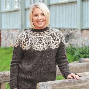 May include: A dark brown sweater with a cream-colored floral pattern around the neckline and shoulders. The sweater has long sleeves and a crew neck. The woman is smiling and standing near a wooden fence.