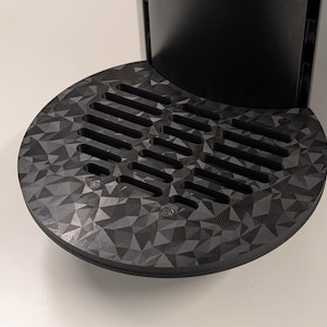 May include: Close-up of a coffee machine's drip tray. The tray is round, black, and features a geometric pattern. It has a series of parallel slots for drainage. The machine's base is dark gray.