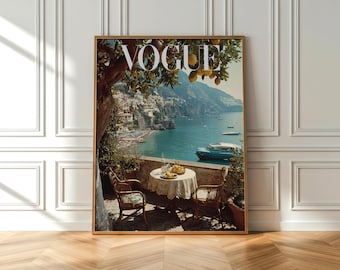 Op de afbeelding: Een ingelijste print van een vintage Vogue-cover met een kustlandschap met een tafel voor twee personen. De afbeelding bevat het woord "VOGUE" in grote witte letters, een blauwe zee en een stad op een heuvel.