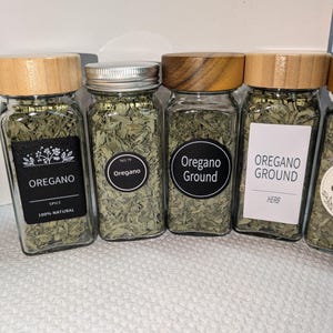 May include: Four rectangular glass spice jars filled with dried green oregano leaves. The jars feature wooden or metal lids and various labels with text including OREGANO, NO.79, Oregano Ground, and HERB. The jars are arranged in a row.