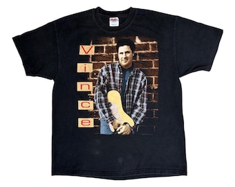 May include: A black short-sleeve t-shirt featuring a graphic print of a person wearing a plaid shirt and holding a guitar against a brick wall background. The name "Vince" is printed vertically in red letters on the left side of the graphic.