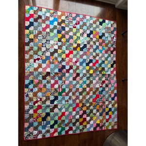 Op de afbeelding: Een rechthoekige patchwork quilt met een herhalend patroon van in elkaar grijpende stoffen vierkanten in een grote verscheidenheid aan kleuren, waaronder rood, blauw, geel, groen en bruin, met diverse patronen zoals ruitjes, bloemen en effen prints, afgewerkt met een rode rand.