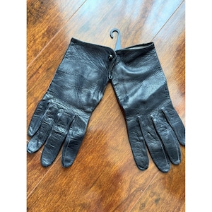 Könnte beinhalten: Ein Paar schwarze Lederhandschuhe mit strukturierter Oberfläche, genähten Fingerdetails und einem kleinen schwarzen Knopfverschluss am Handgelenk. Die Handschuhe sind durch einen schwarzen Kunststoff-Aufhänger zwischen den Öffnungen verbunden.