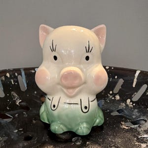 May include: A ceramic pig figurine. It has a white body, pink ears, cheeks, and snout, and black eyes. The pig is wearing green overalls. The figurine has a vintage, cartoonish style.
