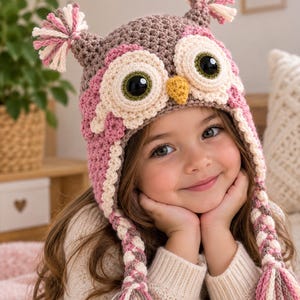 May include: A crocheted owl hat in shades of brown, pink, and cream. The hat features two large circular eyes with black pupils, a small yellow beak, and two ear flaps with braided tassels. Two additional tassels sit atop the hat.