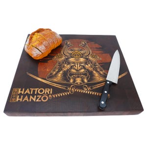 Può includere: Un tagliere rettangolare in legno con un disegno di guerriero samurai, una pagnotta di pane e un coltello. La tavola presenta la scritta "HATTORI HANZO" ed è realizzata in legno scuro con accenti di legno più chiaro. Il coltello ha un manico nero.