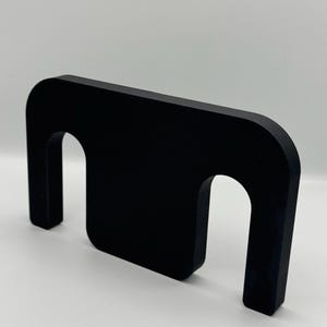 May include: A solid black, geometric object with a rectangular top and two arched openings. The object is made of a matte material and stands upright against a white background. The design is minimalist and modern.