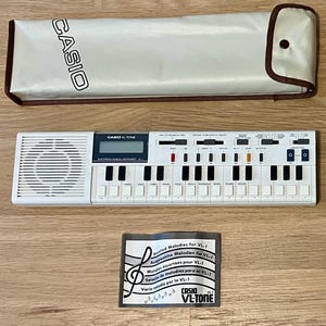May include: A white Casio VL-1 electronic musical instrument with black keys and a small LCD screen. Includes a beige carrying case with the Casio logo and a small instruction card. The instrument is approximately 12 inches long.