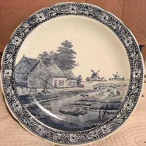 May include: A round, white ceramic plate with a blue and white pastoral scene. The plate features a detailed illustration of a rural landscape with buildings, windmills, a river, and a decorative floral border. The plate has a scalloped edge.