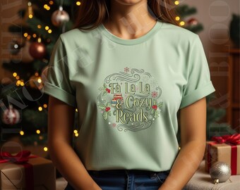 Op de afbeelding: Een lichtgroen T-shirt met opgerolde mouwen met de tekst "Fa La La & Cozy Reads" in een decoratief ontwerp. Het ontwerp bevat boeken, hulst en een sneeuwvlok. Het shirt wordt gecombineerd met een blauwe jeans.