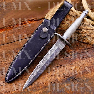 May include: A Damascus steel dagger with a textured silver handle and a black leather sheath. The blade has a distinctive pattern, and the sheath features a snap closure. The dagger is on a wooden surface.