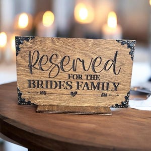 May include: A wooden sign with the words "Reserved for the Brides Family" in elegant script. The sign is brown with decorative black corner accents and sits on a small wooden stand. A heart and arrow design is at the bottom.