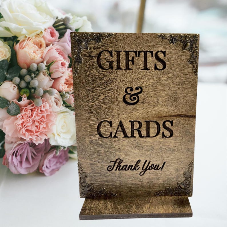 May include: A wooden sign with the words "GIFTS & CARDS" engraved in a dark font. The sign has decorative corner accents and the words "Thank You!" at the bottom. The sign is placed on a white surface with a bouquet of flowers in the background.