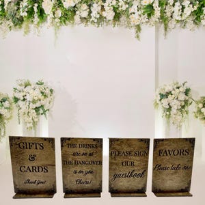 May include: Four wooden wedding signs with decorative borders. The signs read "Gifts & Cards", "The drinks are on us, the hangover is on you, Cheers!", "Please sign our guestbook", and "Favors, Please take one". The signs are set against a white backdrop with floral arrangements.
