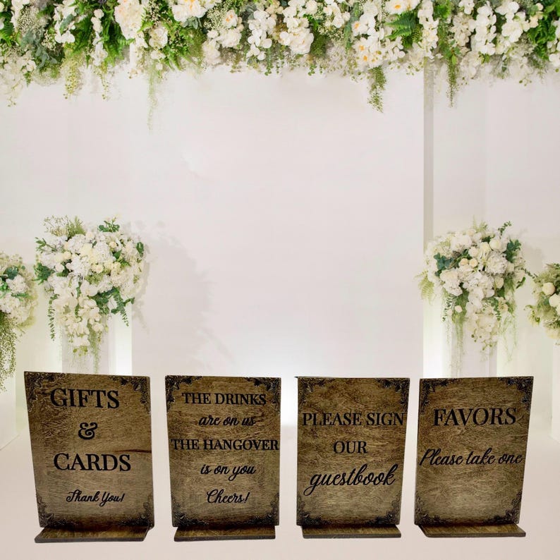 May include: Four wooden wedding signs with decorative borders. The signs read "Gifts & Cards", "The drinks are on us, the hangover is on you, Cheers!", "Please sign our guestbook", and "Favors, Please take one". The signs are set against a white backdrop with floral arrangements.