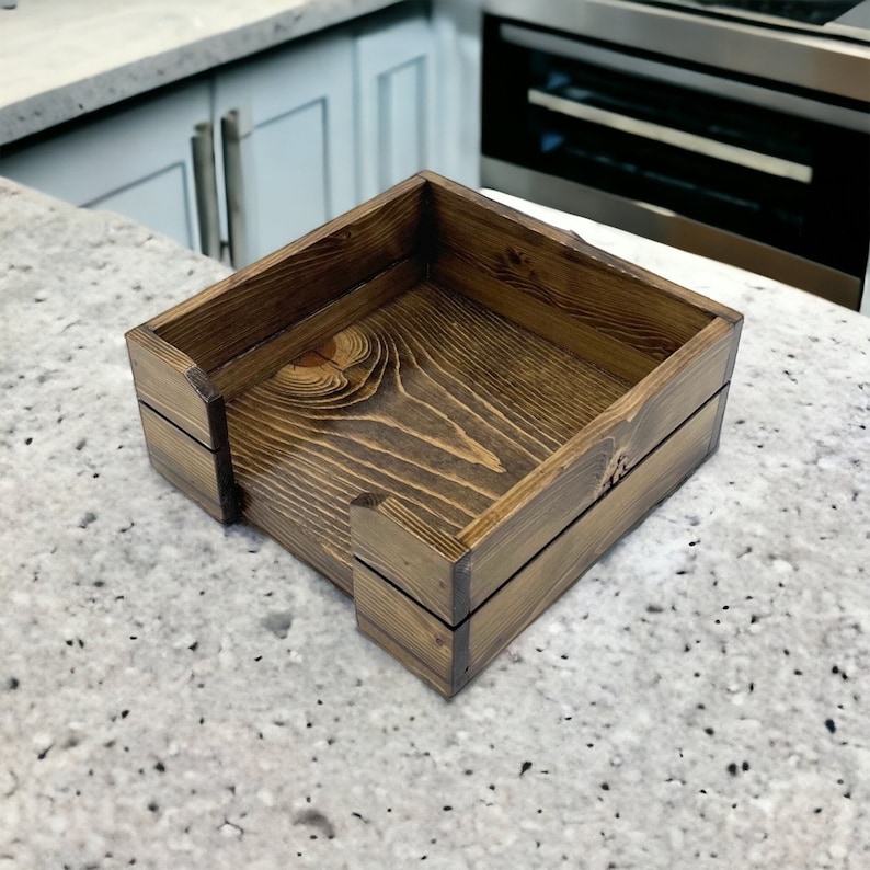 May include: A wooden napkin holder with a dark brown stain. The holder is made of natural wood with visible grain and is square shaped.