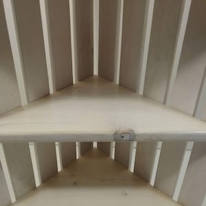 May include: White corner shelf unit with three shelves. The shelves are made of wood and have a slatted design.