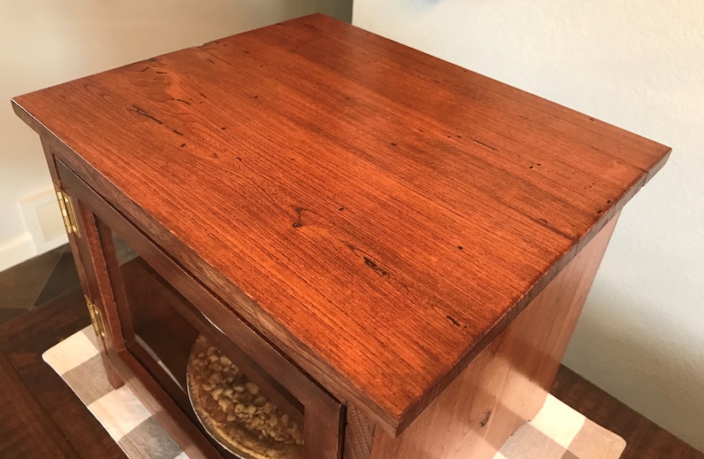 May include: A rectangular wooden cabinet with a rich reddish-brown finish and a visible wood grain. The cabinet features a flat top surface and a glass-paneled door with brass-colored hinges, revealing a circular pie dish stored inside.
