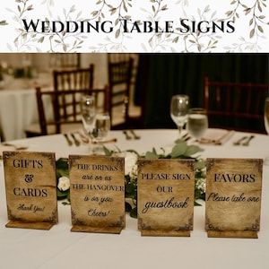 May include: Four wooden wedding table signs with text: "GIFTS & CARDS Thank You!", "THE DRINKS are on us THE HANGOVER is on you Cheers!", "PLEASE SIGN our guestbook", and "FAVORS Please take one". The signs are brown with decorative borders.