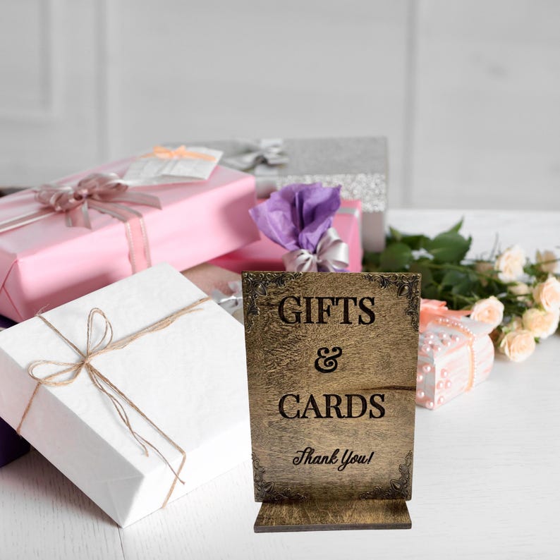 May include: A wooden sign with the words "GIFTS & CARDS Thank You!" in a decorative font. The sign is brown with ornate corner designs. Gift boxes wrapped in pink, white, and silver are in the background.