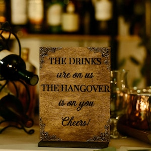 May include: A rectangular wooden sign with decorative corner accents. The sign reads "THE DRINKS are on us THE HANGOVER is on you Cheers!" in black lettering. The sign is on a table with a wine rack and bottles in the background.