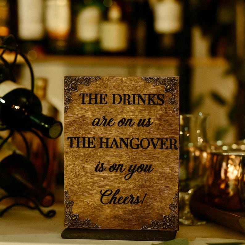 May include: A rectangular wooden sign with decorative corner accents. The sign reads "THE DRINKS are on us THE HANGOVER is on you Cheers!" in black lettering. The sign is on a table with a wine rack and bottles in the background.