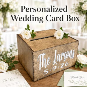 May include: A rectangular wooden card box with a dark wood grain finish, featuring a horizontal slot on top. The front is personalized with the white text The Larsons and the date 5-9-26. Two white roses are placed on the top lid of the box.