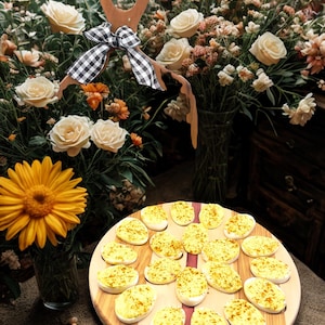 May include: A wooden serving board with deviled eggs, a decorative wooden spoon with a black and white checkered bow, and a floral arrangement with yellow and white flowers.