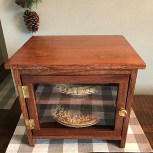 May include: A rectangular wooden display cabinet with a rich brown finish, featuring a glass-front door with a gold-toned knob and two brass hinges. Inside, two shelves hold round pies in metal tins. The cabinet has a flat top and a sturdy frame.