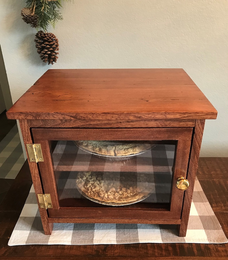 May include: A rectangular wooden display cabinet with a rich brown finish, featuring a glass-front door with a gold-toned knob and two brass hinges. Inside, two shelves hold round pies in metal tins. The cabinet has a flat top and a sturdy frame.