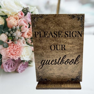 May include: A wooden sign with the text "PLEASE SIGN OUR guestbook" in a decorative font. The sign is brown with ornate corner designs. A bouquet of flowers is in the background.