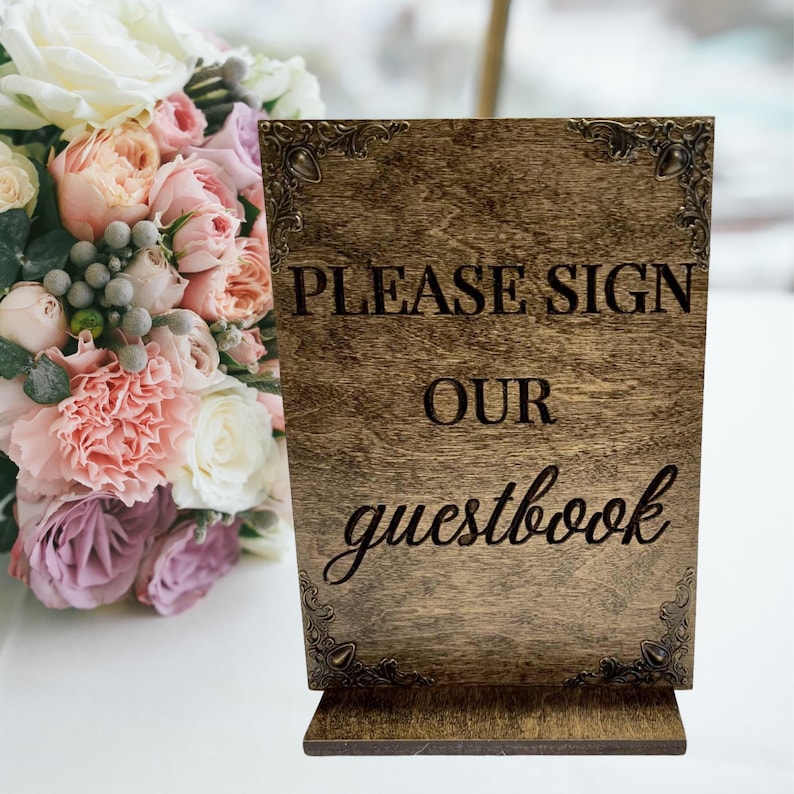 May include: A wooden sign with the text "PLEASE SIGN OUR guestbook" in a decorative font. The sign is brown with ornate corner designs. A bouquet of flowers is in the background.