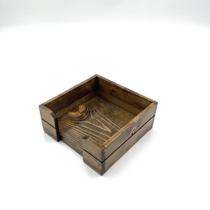 May include: A square wooden napkin holder with a dark brown stain. The holder has a simple design with a slightly raised lip around the top edge.