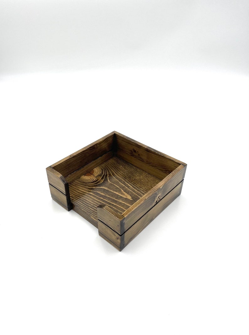 May include: A square wooden napkin holder with a dark brown stain. The holder has a simple design with a slightly raised lip around the top edge.