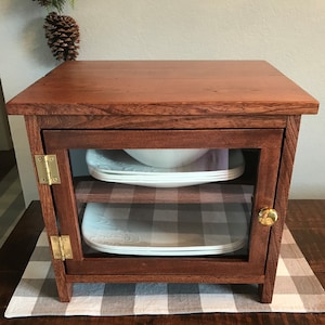 May include: A small, rectangular wooden cabinet with a rich brown finish, featuring a single glass-paneled door with brass hinges and a round brass knob. Inside, two white square plates with embossed edges are visible on the wooden shelf.
