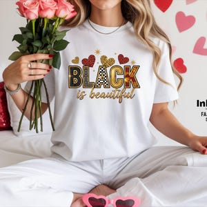 May include: White t-shirt featuring the text "BLACK is beautiful" in a decorative font, with heart and star accents. The shirt has a crew neck and short sleeves. The image also includes pink roses.