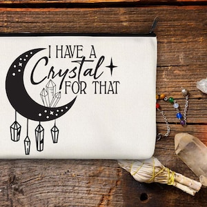 Puede incluir: Una bolsa de lona blanca con cremallera negra, con el texto "I HAVE A Crystal FOR THAT" y un diseño de media luna con cristales. Varios cristales y un manojo de salvia están dispuestos sobre una superficie de madera.