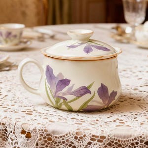 May include: Cream-colored ceramic mug with a lid, adorned with purple iris flower illustrations. The mug has a curved handle and a scalloped rim. The lid also features an iris design. The mug is set on a white lace tablecloth.