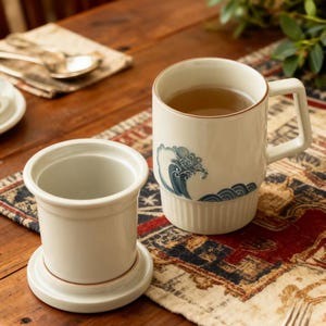 May include: Cream-colored ceramic mug with a blue wave design, filled with tea. The mug has a square handle and a ribbed base. Beside it is a matching tea infuser with a saucer. The items are on a patterned table runner.