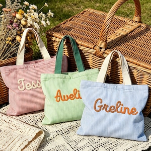Könnte beinhalten: Drei gestreifte Tragetaschen in Rot, Grün und Blau, jede mit einem anderen Namen bestickt: Jess, Aveli und Grelin. Die Taschen haben cremefarbene Griffe. Sie liegen auf einem weißen Spitzentuch, mit einem Weidenkorb im Hintergrund.