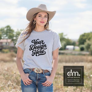 Op de afbeelding: Lichtgrijs t-shirt met de tekst "Your Design Here" in zwart script. De persoon draagt een cowboyhoed, een blauwe spijkerbroek en een decoratieve gesp. De achtergrond is een veld met bomen en een bewolkte lucht.