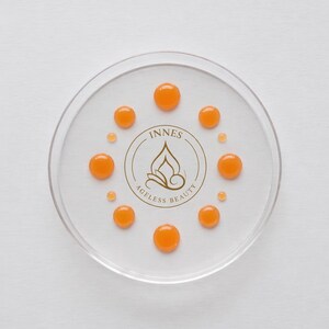 May include: A clear, circular dish with several orange droplets arranged around a central logo. The logo reads "INNES AGELESS BEAUTY" in a gold color. The droplets are evenly spaced, creating a visually appealing pattern.