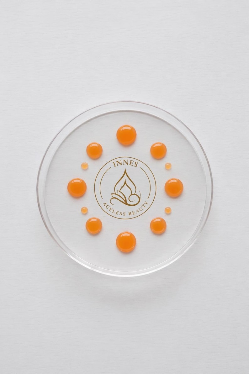 May include: A clear, circular dish with several orange droplets arranged around a central logo. The logo reads "INNES AGELESS BEAUTY" in a gold color. The droplets are evenly spaced, creating a visually appealing pattern.