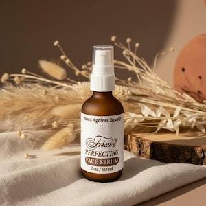 Pu&ograve; includere: Una bottiglia di vetro marrone di Firm Perfecting Face Serum con un dosatore a pompa bianco. L'etichetta recita "Innes Ageless Beauty" e "2 oz/60 ml". La bottiglia &egrave; appoggiata su un panno colorato neutro con piante secche sullo sfondo.