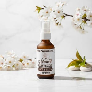 Pu&ograve; includere: Una bottiglia di vetro marrone di Firm Perfecting Face Serum, 60 ml, con pompa ed etichetta bianche. L'etichetta include il testo "Innes Ageless Beauty". Lo sfondo presenta fiori bianchi e una superficie bianca.